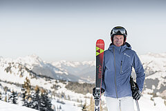 Person standing with skis in a winter alpine landscape, snow-covered mountains visible in the background.