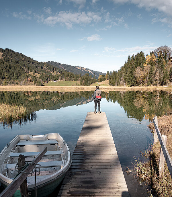 Mann schaut vom zuvorderst beim Steg über den herbstlichen See hinaus, neben dem Steg ruht ein Ruderboot. Hintergrund mit waldbedeckten Bergen.