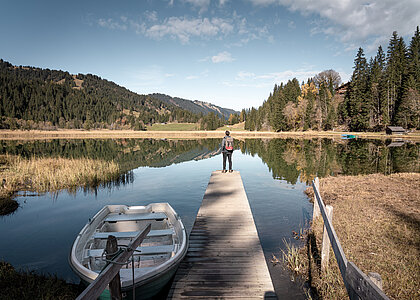 Mann schaut vom zuvorderst beim Steg über den herbstlichen See hinaus, neben dem Steg ruht ein Ruderboot. Hintergrund mit waldbedeckten Bergen.