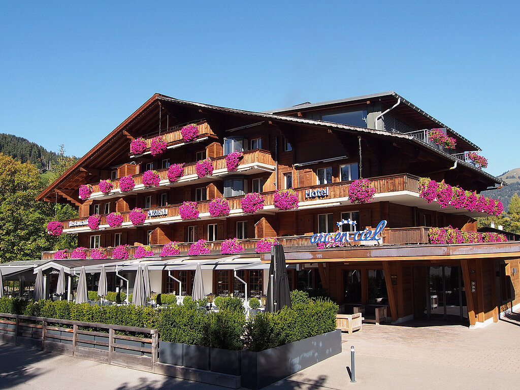 Sicht auf das mit Blumen geschmückte Hotel Arc-en-ciel, im Vordergrund hinter grünen Pflanzen ist die Terrasse ersichtlich.