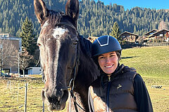 Person standing outdoors next to a horse on a grassy field; buildings, trees and forested mountain slopes visible in the background.