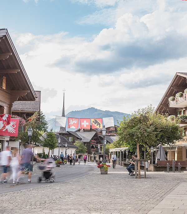 Promenade bordée de chalets fleuris, d’une chapelle et de lampadaires sur laquelle des piétons déambulent.