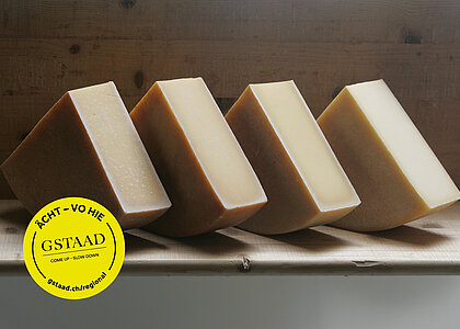 Auf einem Holzbrett stehen viermal ein Viertel eines Hartkäses. Die Wand dahinter ist auch aus Holz. Auf der linken unteren Seite ist ein gelber, runder Kleber platziert. Darauf steht Gstaad - Come up - Slow down in der Mitte und am Rand jeweils ÄCHT - VO HIE und gstaad.ch/regional