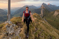 Hiker walking on a narrow mountain ridge with a summit cross and panoramic Alpine views.