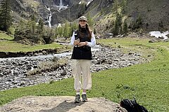 Centrally positioned person standing on a rock in an alpine landscape with a stream, green meadows, conifer trees and steep mountain slopes in the background.