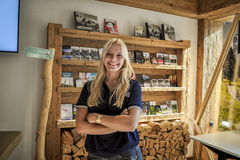 Mitarbeitende von Gstaad Saanenland Tourismus steht vor einem rustikalen Holzregal mit Broschüren und Informationsmaterial im Tourismusbüro Gstaad.