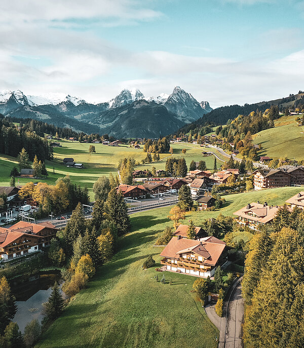 Aus der Vogelperspektive sieht man das Dorf Saanenmöser im Sommer. Die Flächen rund ums Dorf sind grün und im Hintergrund sieht man höhere Berge.