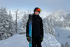 Personne debout dans la neige avec des skis sur une arête de montagne, entourée d’arbres enneigés et d’un paysage alpin.