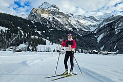 Person standing on cross-country skis on a prepared trail, with snow-covered mountains and an Alpine village behind.