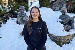Person steht zentral im Bild im Freien auf schneebedecktem Boden. Im Hintergrund sind mehrere Skulpturen von musizierenden Figuren zu sehen, die teilweise von Schnee bedeckt sind. Steine, Pflanzen und Schnee prägen die Umgebung.