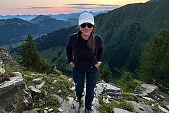 Personne debout sur une crête rocheuse avec vue sur les pentes alpines verdoyantes au coucher du soleil.
