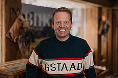 Personne debout dans un intérieur rustique en bois, portant un pull avec l’inscription « GSTAAD », décoration alpine visible en arrière‑plan.