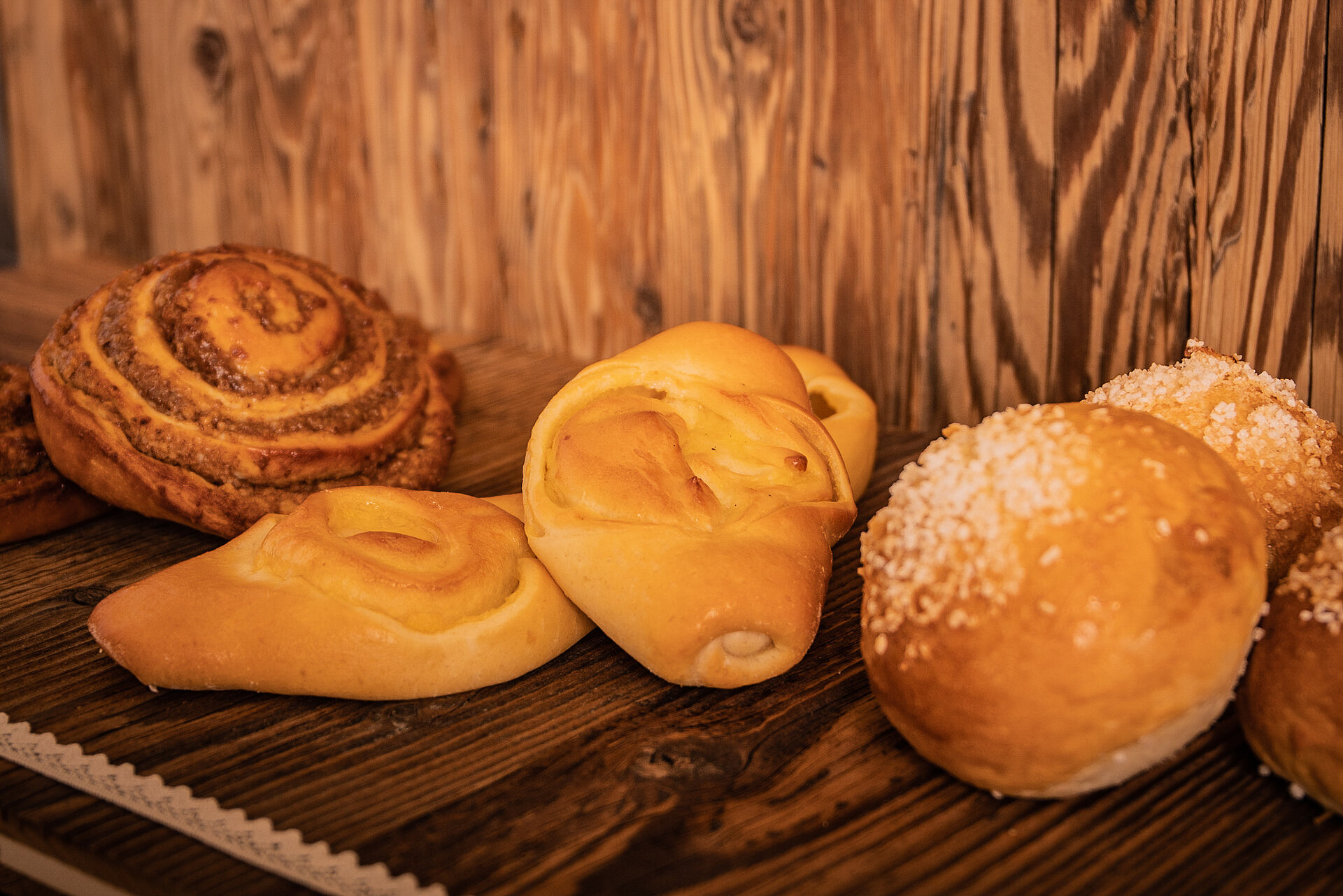 Auf einem braunen Holzgestell liegen verschiedenen Patisserie. Links steht eine Nuss-Schnecke, gefolgt von zwei Vanille-Gipfeln und drei Berliner mit Zuckerguss.