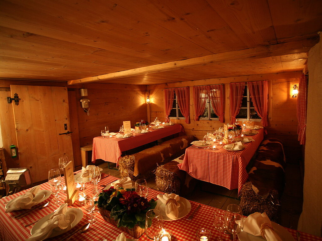 <p>Ein rustikaler Raum mit rot-weiss karierten Tischen, gedeckt für ein Abendessen. Bänke aus Strohballen mit Kuhfellen.</p>
