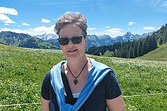 Person wearing sunglasses on a green alpine meadow filled with wildflowers. Forested slopes, snow-capped mountain peaks and a clear blue sky can be seen in the background.