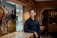 Personne debout dans un intérieur rustique en bois, à côté d’une vitrine d’exposition ; décoration alpine et panneaux d’information visibles en arrière‑plan.