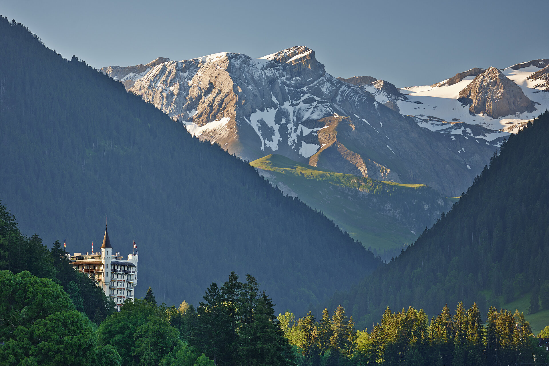 Das Hotel Gstaad Palace ist gebaut wie ein Schloss und überragt den Wald. Im Hintergrund sieht man Schneeberge die von der Morgensonne bestrahlt werden