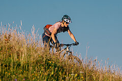 Person riding a mountain bike downhill across a grassy slope, wearing a helmet and backpack; blue sky in the background.