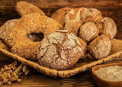Auf einem Holzgestell liegt ein geflochtener viereckiger Korb. Darin ist ein Roggenbrot und ein dunkler Brotkranz mit Sesam und Kernen drauf. Rechts daneben ist ein dunkles, aus sechs kleinen Brötchen geformtes Brot. Im Hintergrund sind weitere vier dunklere Brote zu sehen. Im Vordergrund sind getrocknete Weizen- und Roggenähren zu sehen, sowie eine kleine Schüssel mit Mehl.