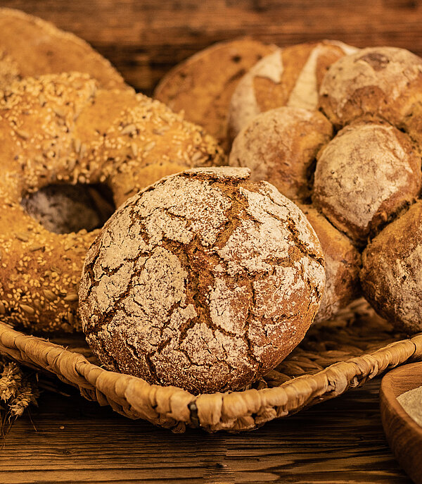 Auf einem Holzgestell liegt ein geflochtener viereckiger Korb. Darin ist ein Roggenbrot und ein dunkler Brotkranz mit Sesam und Kernen drauf. Rechts daneben ist ein dunkles, aus sechs kleinen Brötchen geformtes Brot. Im Hintergrund sind weitere vier dunklere Brote zu sehen. Im Vordergrund sind getrocknete Weizen- und Roggenähren zu sehen, sowie eine kleine Schüssel mit Mehl.