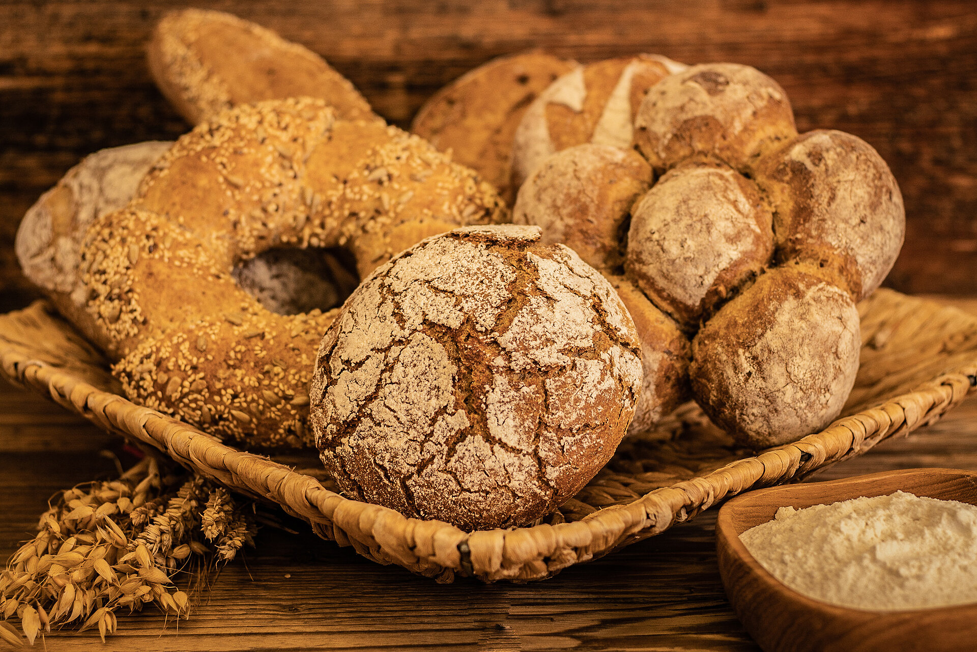 Auf einem Holzgestell liegt ein geflochtener viereckiger Korb. Darin ist ein Roggenbrot und ein dunkler Brotkranz mit Sesam und Kernen drauf. Rechts daneben ist ein dunkles, aus sechs kleinen Brötchen geformtes Brot. Im Hintergrund sind weitere vier dunklere Brote zu sehen. Im Vordergrund sind getrocknete Weizen- und Roggenähren zu sehen, sowie eine kleine Schüssel mit Mehl.