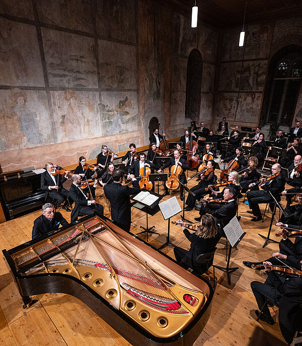 An orchestra performs classical music in a church, with Christian motifs visible on the walls.