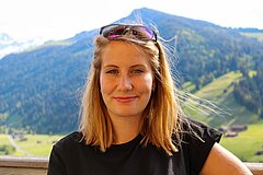 Portrait of a person with sunglasses on their head, standing in front of green mountains.