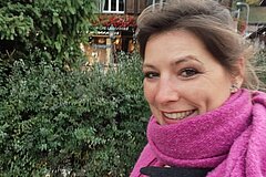 Close-up of a person outdoors. The face is visible in the foreground, and the person is wearing a scarf. In the background, green shrubs and a wooden building with windows and decorative elements can be seen.