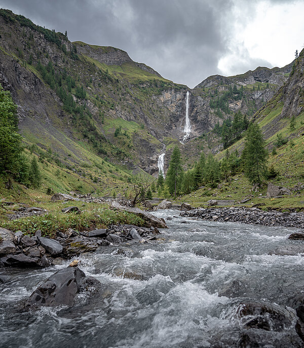 An impressive waterfall cascades down into the valley. There is a stream in the foreground, with mountains behind it.