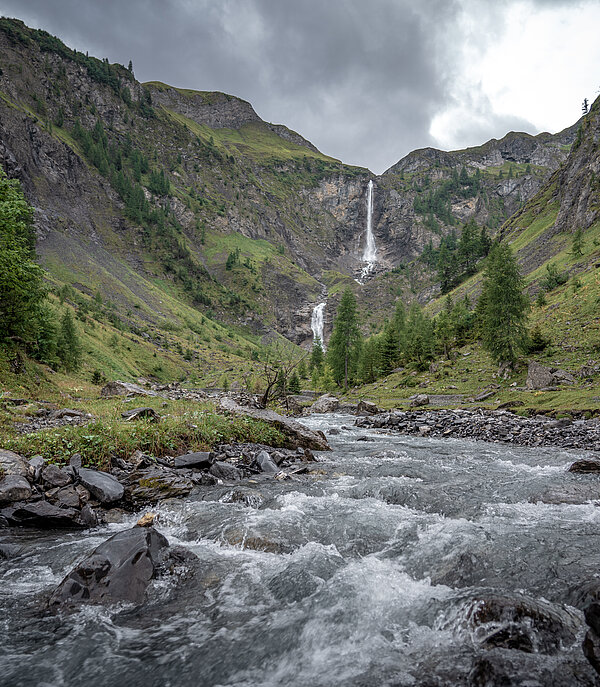 Ein imposanter Wasserfall stürzt sich ins Tal hinunter. Im Vordergrund ein Bach, dahinter Berge.