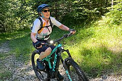 Personne portant un casque faisant du VTT sur un chemin de gravier à travers une forêt ensoleillée.