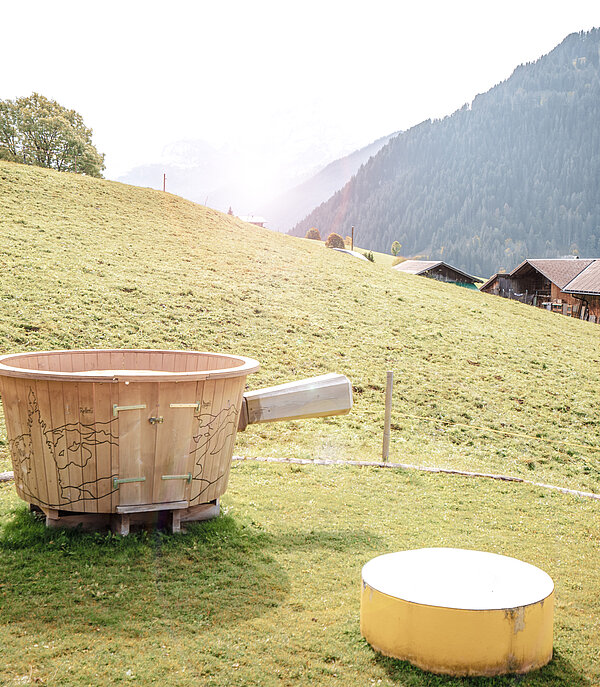Oversized fondue caquelon to sit on stands on a lawn. In the background you can see a wooden barn and the Wispile mountain.