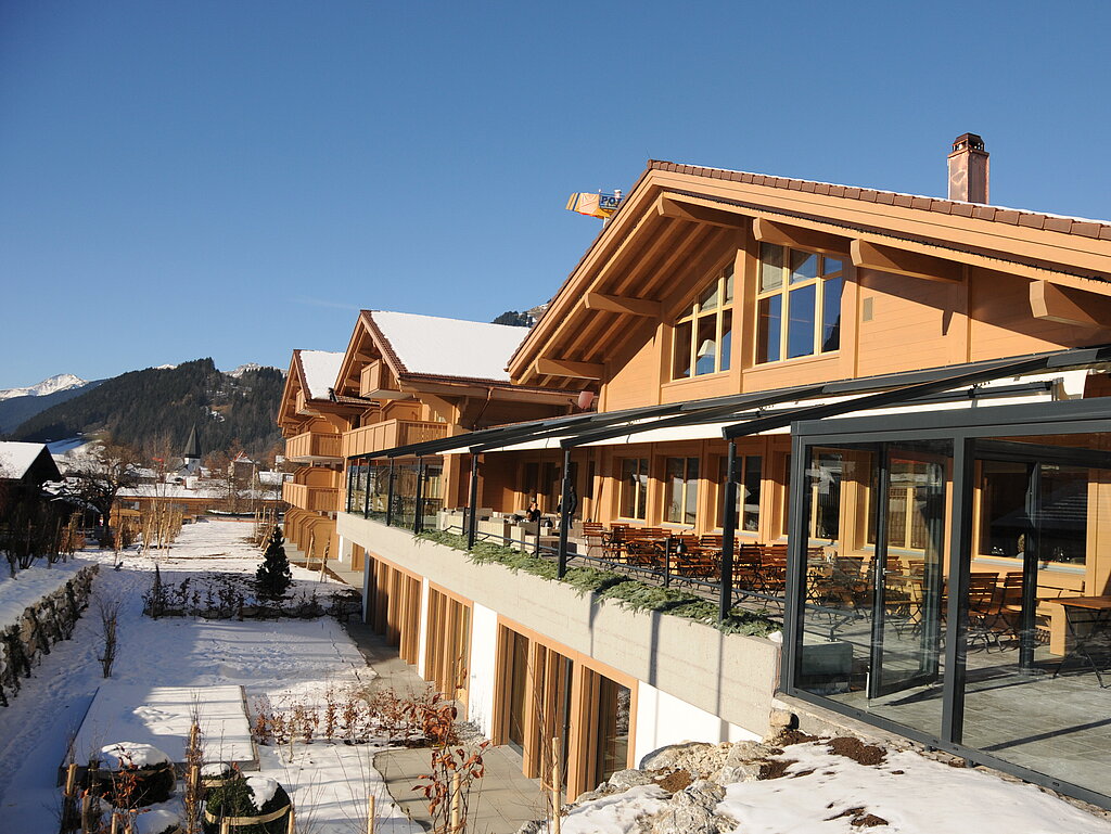 Moderne Gebäude im Chaletstil mit grossen Fenstern und sonnigen Terrassen vor schneebedeckter Bergkulisse.