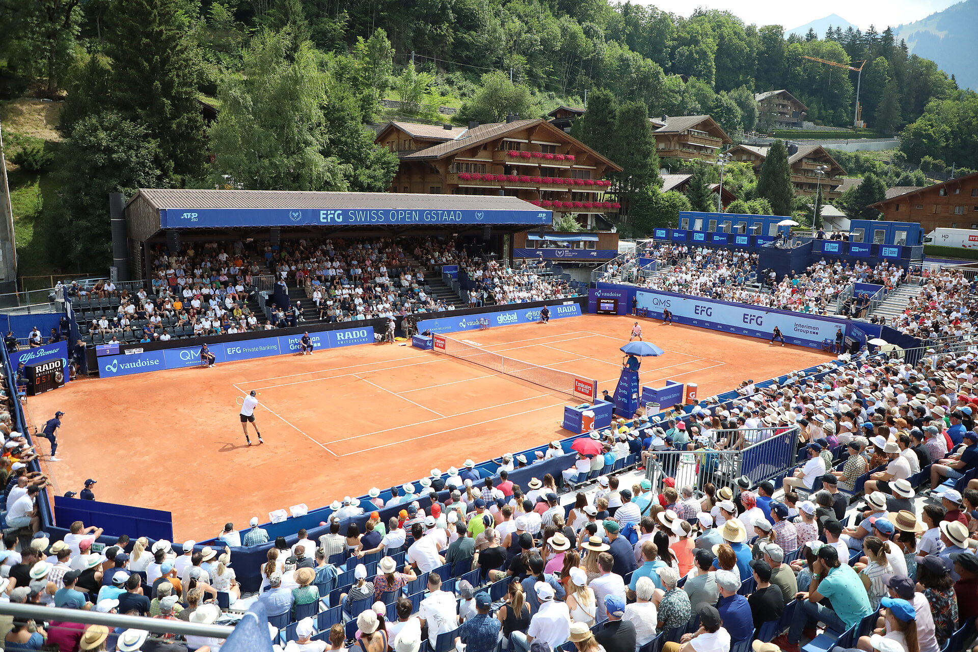 Tennisspiel mit Spielern und Publikum in Gstaad, im Hintergrund Wald und Häuser.