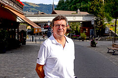 Mitarbeiter in weissem Poloshirt steht auf einer Dorfstrasse vor Gebäuden und Bergkulisse, im Hintergrund sind Bäume, Sitzbänke und ein Kirchturm sichtbar.