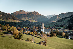 Blick auf ein Dorf mit einer grossen Kirche, mit Bergen und farbigen Bäumen im Hintergrund.