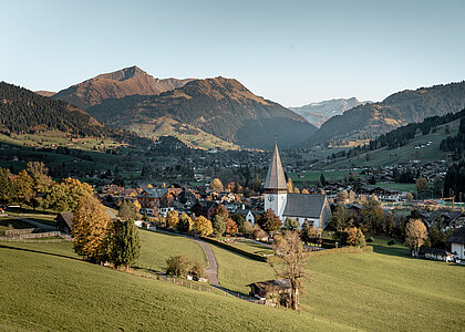 Blick auf ein Dorf mit einer grossen Kirche, mit Bergen und farbigen Bäumen im Hintergrund.