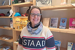 Person standing indoors in front of light wooden shelves. The shelves contain brochures, books, hats and small souvenir items. The person is wearing a light-colored sweater and a scarf and is positioned centrally in the image.
