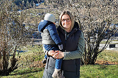 Adult holding a child outdoors; they are standing on a grassy area with bushes, landscape and buildings visible in the background.