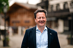 Person standing outdoors in front of traditional wooden buildings in an alpine village, background slightly blurred.