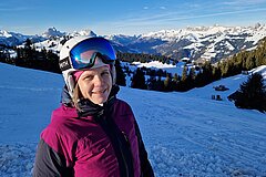Person wearing a ski helmet and goggles standing on a ski slope with snow-covered mountains in the background.