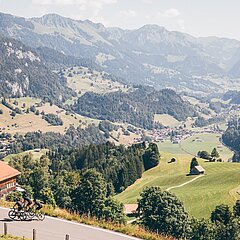 Drei Männer fahren auf einer Passstrasse gegen den Pass. Im Hintergrund sieht man das Simmental im Hochsommer mit Bäumen, Wiesen und Wäldern.