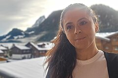 Portrait d’une personne au premier plan, avec des toits enneigés et des montagnes dans un paysage alpin hivernal.