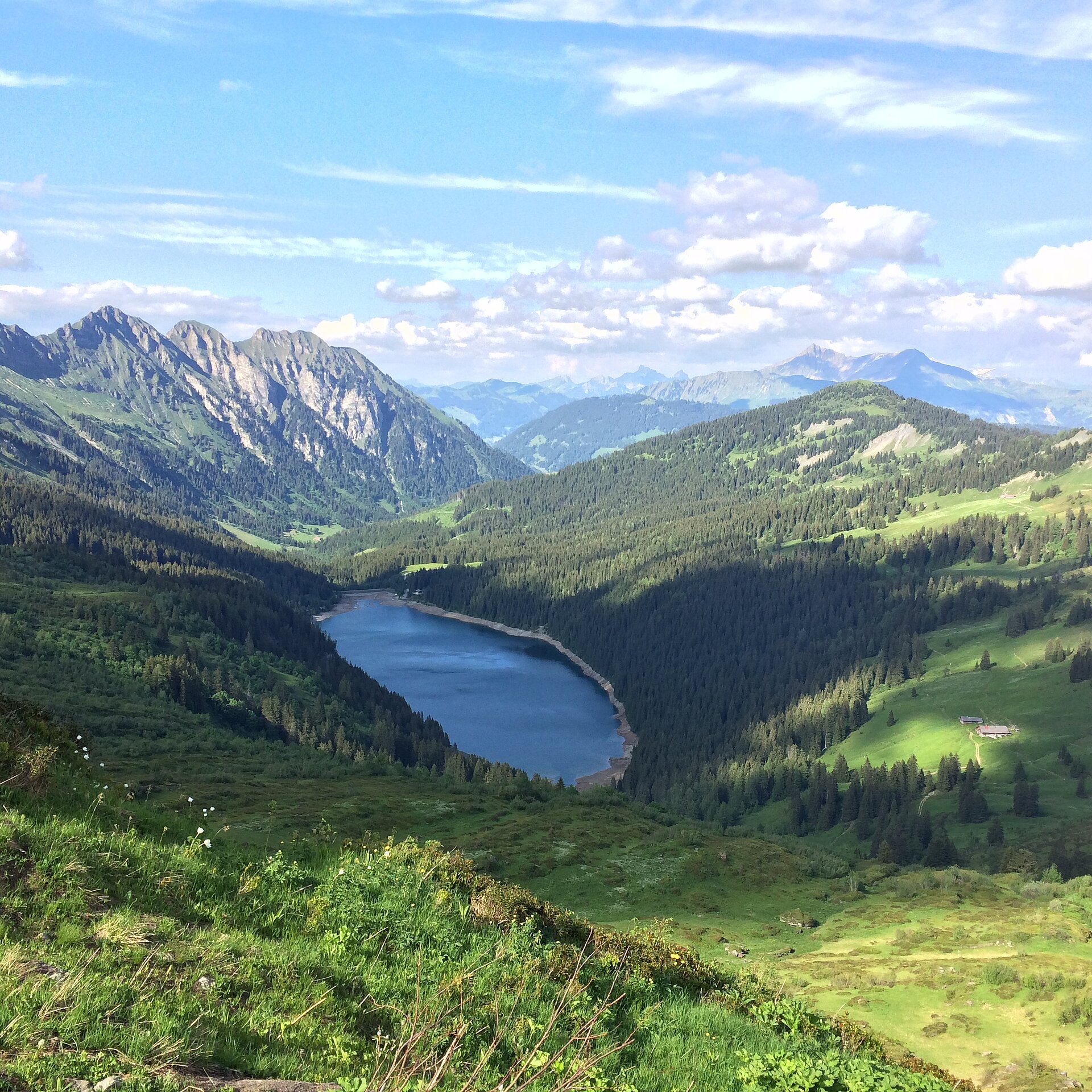 Von einer grasbewachsenen Bergspitzen sieht man tief im Tal einen blauschimmernden Bergsee. Rund herum hat es grüne Weiden und Wald. Hinter dem See ragen drei felsige Berge in den Himmel. Der Himmel ist hellblau mit weissen Wolken.