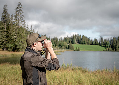Ranger in braun/grüner Kleidung und braunem Cap schaut durch einen Feldstecher. Dahinter sieht man den Lauenensee und darum herum die herbstliche Natur mit Bäumen, Tannen und gelben Gräsern.