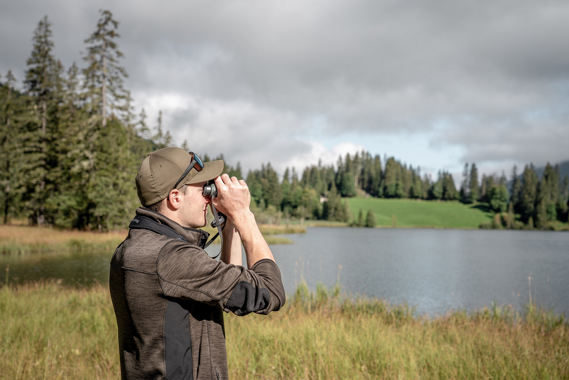 Ranger in braun/grüner Kleidung und braunem Cap schaut durch einen Feldstecher. Dahinter sieht man den Lauenensee und darum herum die herbstliche Natur mit Bäumen, Tannen und gelben Gräsern.