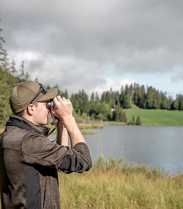 Ranger en tenue marron/verte et casquette marron regardant à travers des jumelles. Derrière, on voit le lac Lauenen et, autour, la nature automnale avec des arbres, des sapins et des herbes jaunes.