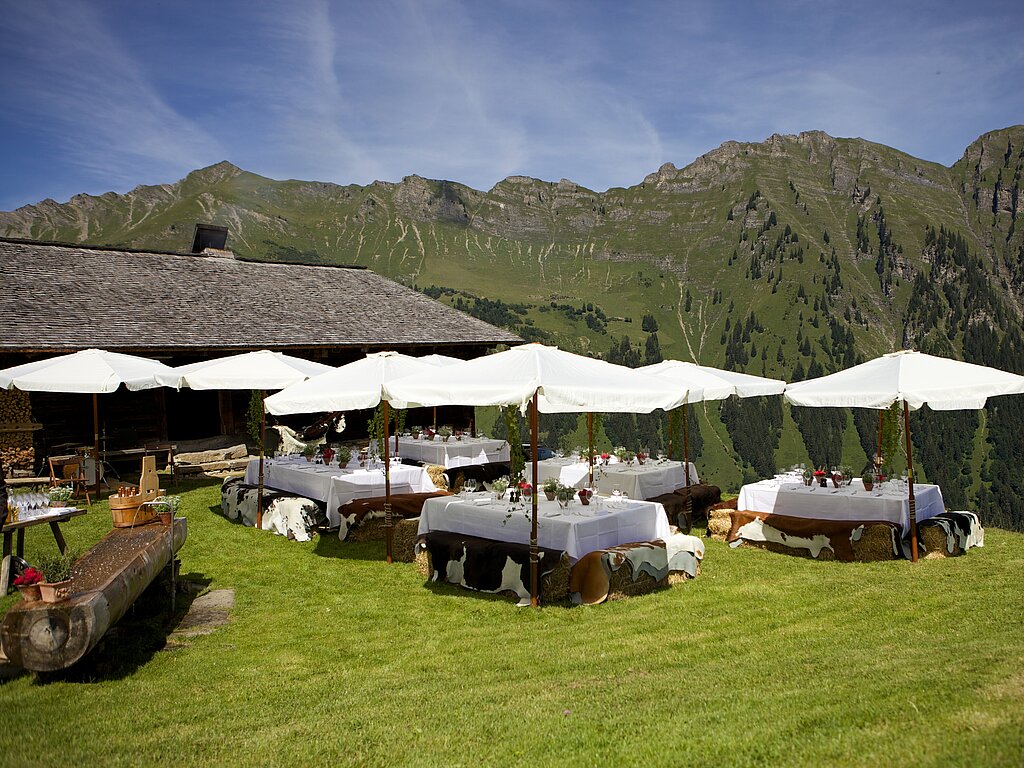 Eine Gruppe von Tischen mit weissen Tüchern und Sonnenschirmen auf einer Wiese, eine Bergkette vor blauem Himmel.