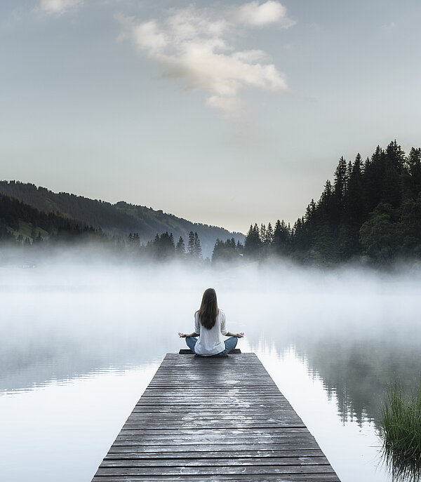 Frau macht Entspannungsübungen auf einem Steg beim See, welcher leicht im Nebel eingehüllt ist.