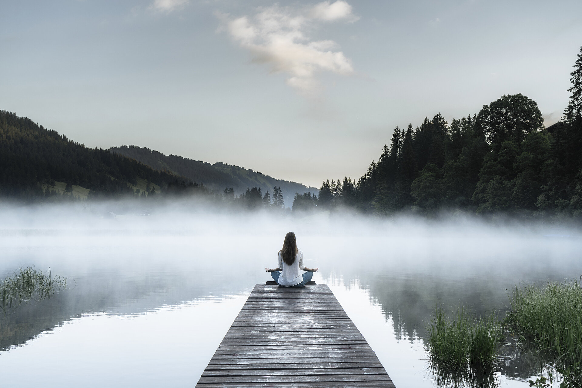 Frau macht Entspannungsübungen auf einem Steg beim See, welcher leicht im Nebel eingehüllt ist.